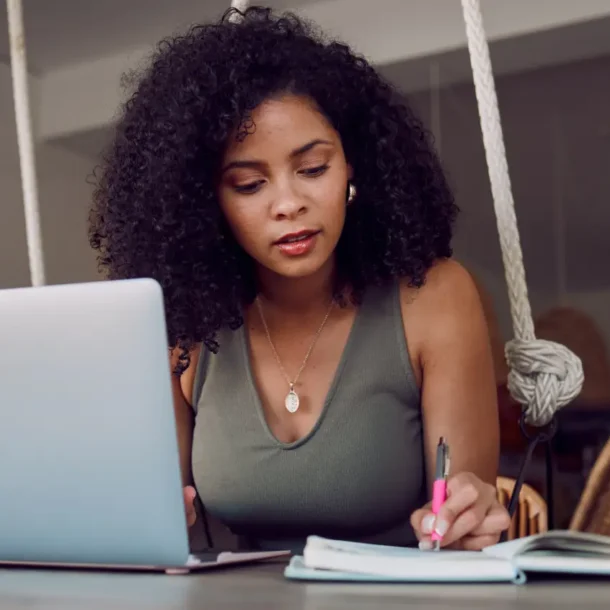 Woman sitting at a laptop, holding a pen, and writing event promotion ideas in a notebook