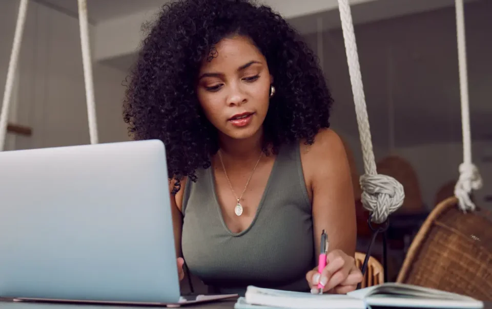 Woman sitting at a laptop, holding a pen, and writing event promotion ideas in a notebook